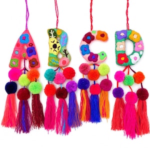 May include: Four hanging fabric letters A, B, C, and D, each embroidered with colourful floral patterns. Each letter features a hanging string and is decorated with multiple vibrant yarn pom-poms and long, dangling tassels in various bright colours.