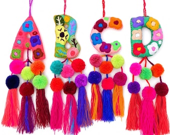 May include: Four hanging fabric letters A, B, C, and D, each embroidered with colorful floral patterns. Each letter features a hanging string and is decorated with multiple vibrant yarn pom-poms and long, dangling tassels in various bright colors.