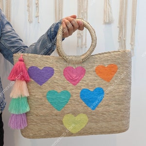 May include: A rectangular woven straw tote bag featuring two round handles and a multi-colored tassel charm. The front of the bag is decorated with six embroidered hearts in purple, pink, orange, teal, blue, and yellow, arranged in a pattern.