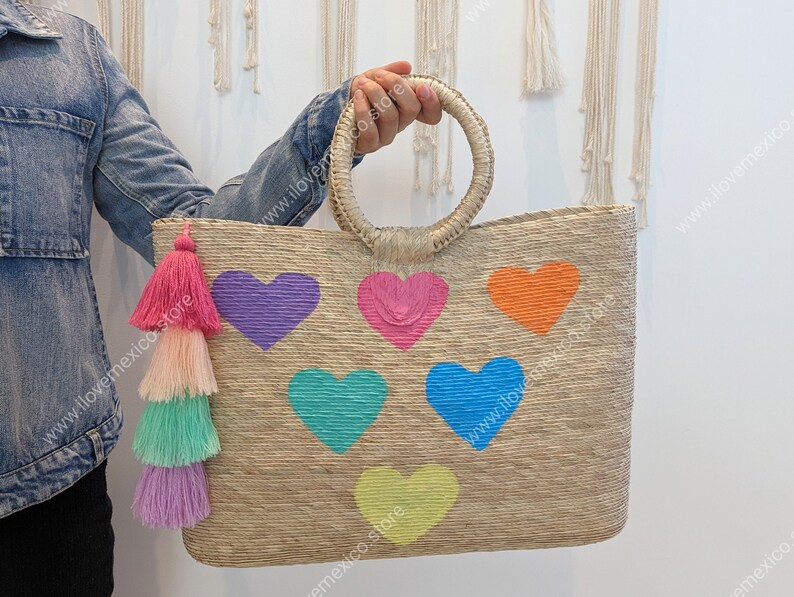 May include: A rectangular woven straw tote bag featuring two round handles and a multi-colored tassel charm. The front of the bag is decorated with six embroidered hearts in purple, pink, orange, teal, blue, and yellow, arranged in a pattern.