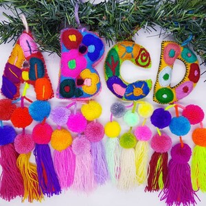 May include: A colourful felt letter garland featuring the letters A, B, C, and D. Each letter is embroidered with floral designs and has pom-pom tassels. This garland would be a lovely addition to a nursery or child's room.