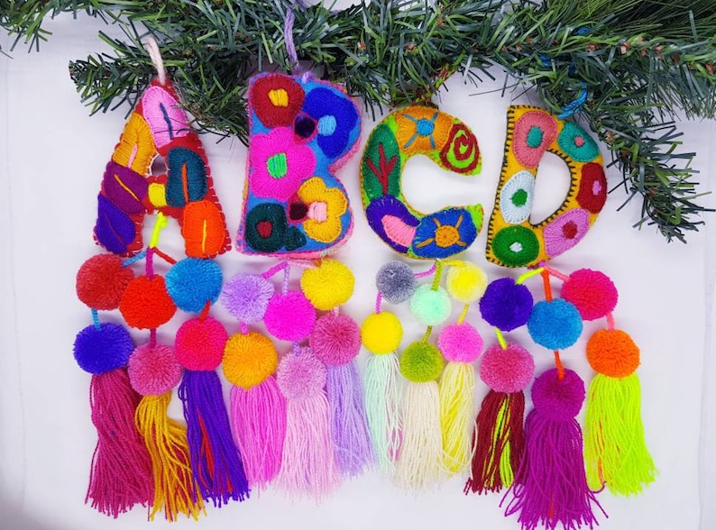 May include: A colourful felt letter garland featuring the letters A, B, C, and D. Each letter is embroidered with floral designs and has pom-pom tassels. This garland would be a lovely addition to a nursery or child's room.