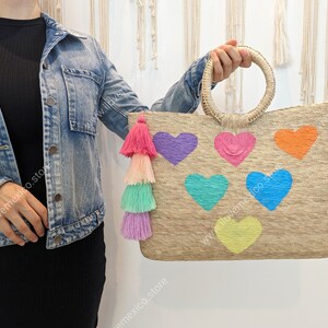 May include: A rectangular woven straw tote bag featuring two round handles and a multi-colored tassel attachment. The front of the bag is decorated with six embroidered heart shapes in purple, pink, orange, teal, blue, and yellow colors.