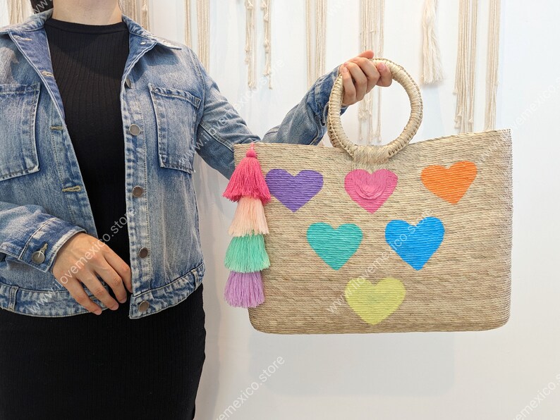 May include: A rectangular woven straw tote bag featuring two round handles and a multi-colored tassel attachment. The front of the bag is decorated with six embroidered heart shapes in purple, pink, orange, teal, blue, and yellow colors.