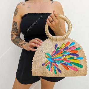 Puede incluir: Un bolso de paja con un diseño de colibrí colorido. El bolso tiene un asa redonda y una forma de semicírculo.