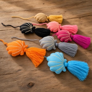 Puede incluir: Un conjunto de seis adornos de tela con forma de concha, cada uno con una borla a juego y un lazo para colgar. Las conchas son de color beige, negro, rosa, magenta, naranja y azul, con detalles de costura en la superficie de cada una.