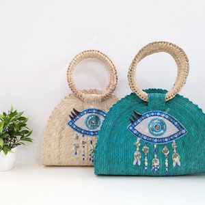 May include: Two straw tote bags with a blue evil eye design. The bags have a half-circle shape and a woven texture. The evil eye design is embellished with sequins and beads.