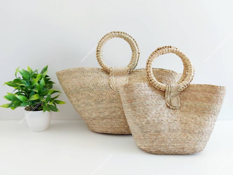 May include: Two natural-colored woven straw bags with round, rigid handles. The bags feature a textured, fibrous surface and a wide, open-top design. The handles are securely attached to the body of each bag with a woven straw tab detail.