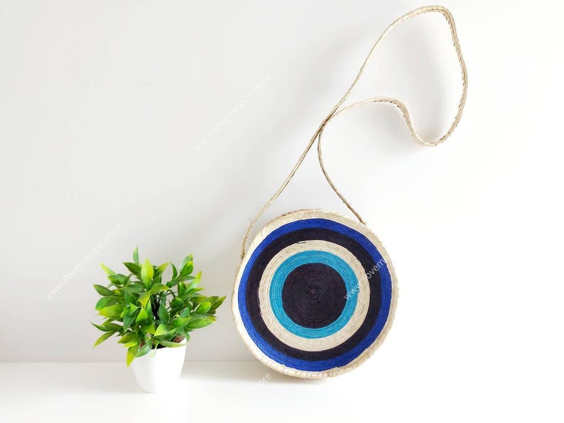 May include: A round, woven bag with a blue, white, and black concentric circle pattern. The bag has a long, woven strap.