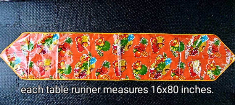 May include: Orange table runner with a yellow border and a repeating pattern of colorful fruit. Each table runner measures 16x80 inches.