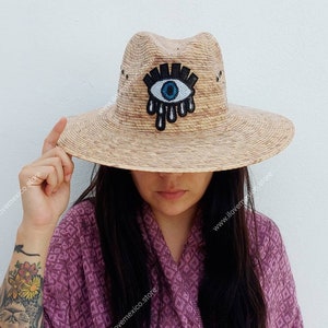 May include: A straw hat with a wide brim and a blue evil eye design. The hat is made of natural straw and has a brown band around the crown.
