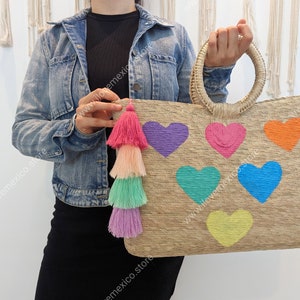 May include: A rectangular woven straw tote bag featuring two round handles and six embroidered heart shapes in purple, pink, orange, teal, blue, and yellow. A multi-layered tassel in shades of pink, peach, mint, and lavender hangs from the side.
