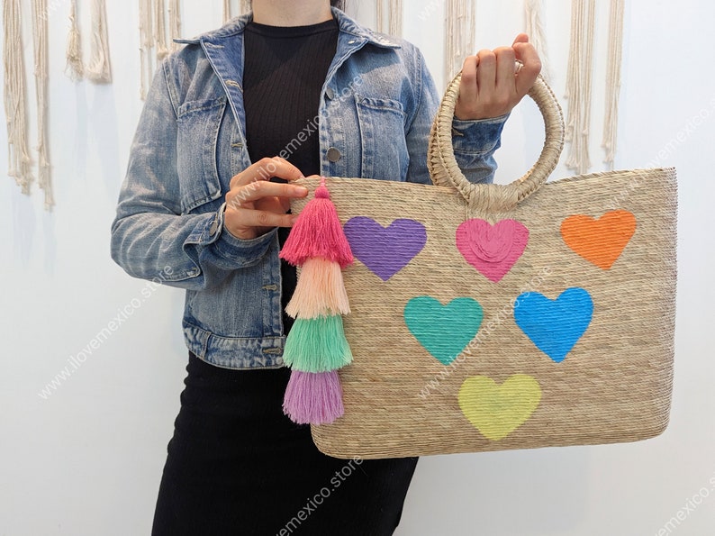 May include: A rectangular woven straw tote bag featuring two round handles and six embroidered heart shapes in purple, pink, orange, teal, blue, and yellow. A multi-layered tassel in shades of pink, peach, mint, and lavender hangs from the side.