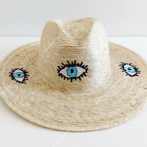 May include: A straw hat with a wide brim and a fedora crown. The hat is decorated with three embroidered blue eyes with white pupils and black eyelashes.