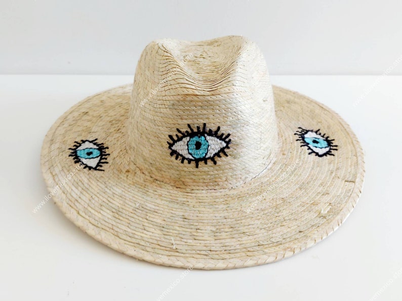 May include: A straw hat with a wide brim and a fedora crown. The hat is decorated with three embroidered blue eyes with white pupils and black eyelashes.
