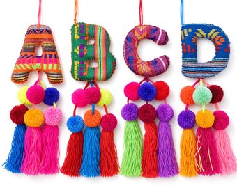 May include: Four hanging fabric letters A, B, C, and D, each featuring vibrant, multi-colored woven patterns. Each letter is suspended by a string and decorated with a cluster of colorful yarn pom-poms and long, dangling tassels in various bright hues.