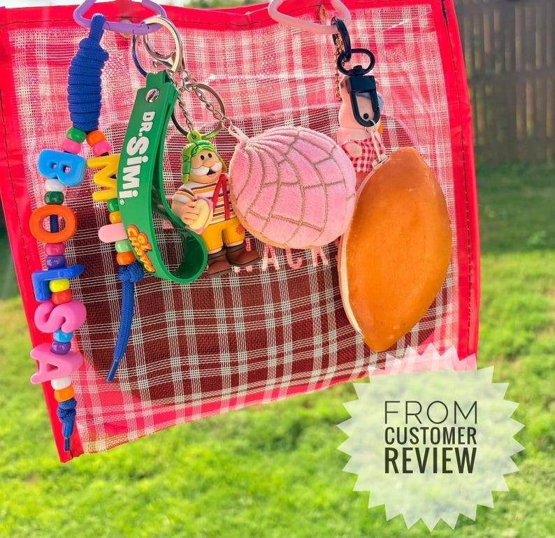 May include: A transparent red and white plaid bag featuring several keychains: a blue, yellow, and pink beaded strand spelling BOLSA, a green Dr. Simi strap, a small figurine of a character in a striped shirt, a pink concha bread charm, and a brown oval charm.
