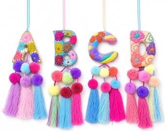 May include: Four hanging ornaments shaped like the letters A, B, C, and D. Each letter is crafted from fabric with colorful floral embroidery and attached to a cluster of small pom-poms and long, vibrant yarn tassels in shades of pink, blue, and yellow.