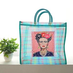May include: A turquoise and blue plaid tote bag with green handles. The bag features a framed image of Frida Kahlo with flowers in her hair. The bag is made of a transparent material.