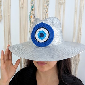 May include: Silver fedora hat with a large, round, blue and white crochet eye design on the front. The hat has a wide brim and a textured appearance. The eye design features a dark blue center, surrounded by white and a larger blue circle.