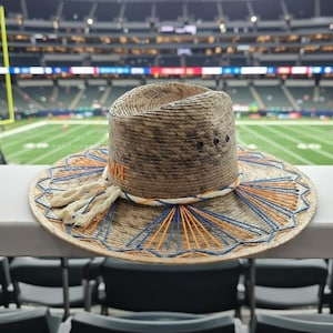 May include: A straw hat with a wide brim, featuring a woven design with blue and orange accents. The hat has a cylindrical crown and is adorned with cream-colored tassels. The background shows a football stadium.