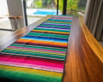 May include: A rectangular table runner featuring a vibrant, multi-colored striped pattern. The textile displays horizontal bands of bright pink, green, yellow, orange, blue, purple, and black, accented with thin white geometric woven details.