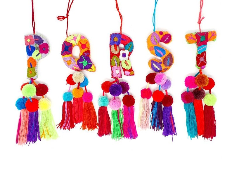 May include: Five colourful felt letters, P, Q, R, S, and T, are embroidered with floral patterns and have pom-poms and tassels hanging from the bottom.