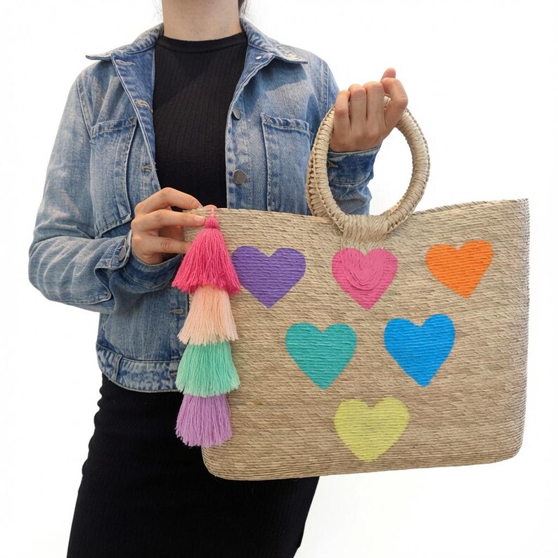 May include: A rectangular woven straw tote bag featuring two round handles and six embroidered hearts in purple, pink, orange, teal, blue, and yellow. A multi-colored tassel in shades of hot pink, peach, mint green, and lavender hangs from the side.
