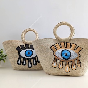 Puede incluir: Dos bolsos de paja con un diseño de ojo turco azul y dorado. Los bolsos tienen una textura de paja tejida y un asa redonda.