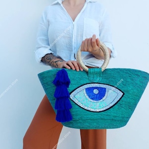 May include: A turquoise woven tote bag with a large blue evil eye design and a blue tassel. The bag has a natural woven handle.