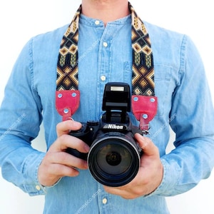 May include: A person wearing a blue denim shirt with a red and black camera strap. The camera strap is woven with a geometric pattern. The person is holding a black camera with a red flash and a silver lens. The camera has the word "Nikon" printed on it.