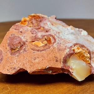 May include: A natural stone specimen with a rough, uneven surface. The stone displays a reddish-brown hue, with lighter cream-coloured inclusions and patches of orange and white. The texture of the stone is clearly visible.