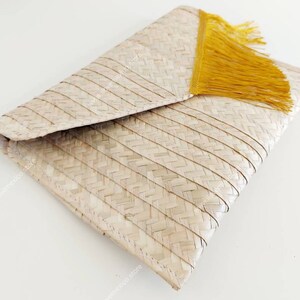 Op de afbeelding: Een geweven strooien envelop-clutch met een lichtbeige natuurlijke vezelstructuur, een visgraatpatroon en een felgele franje aan de diagonale rand van de flap. Het accessoire heeft een gestructureerd, gevouwen ontwerp.