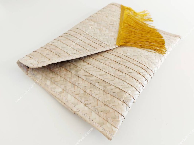 Op de afbeelding: Een geweven strooien envelop-clutch met een lichtbeige natuurlijke vezelstructuur, een visgraatpatroon en een felgele franje aan de diagonale rand van de flap. Het accessoire heeft een gestructureerd, gevouwen ontwerp.