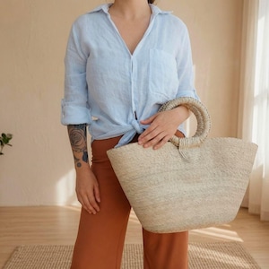 May include: A light blue button-down shirt with long sleeves rolled up and a tied front hem, paired with solid rust-colored trousers. A large, natural-colored woven straw tote bag with two round, braided handles is held in front of the body.