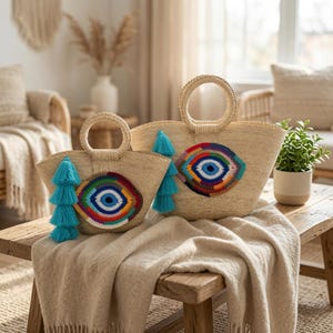 Puede incluir: Dos bolsos de paja trenzada con asas redondas, cada uno con un motivo de ojo turco tejido a ganchillo en azul, verde, amarillo y rojo. Una borla de color turquesa brillante cuelga del lateral de cada bolso, añadiendo un toque de color natural.