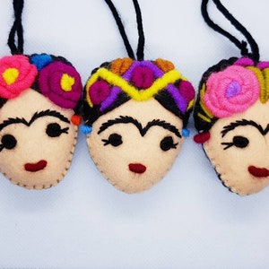 May include: Three felt ornaments shaped like faces with embroidered features. Each face has black hair, thick eyebrows, and a red mouth. They are adorned with colorful yarn flowers or geometric patterns in pink, yellow, purple, and blue, with black hanging cords.