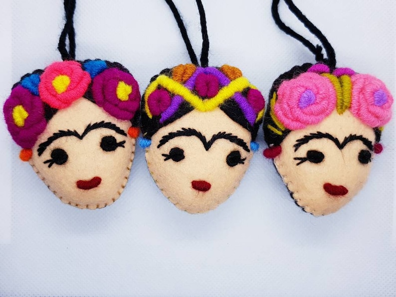 May include: Three felt ornaments shaped like faces with embroidered features. Each face has black hair, thick eyebrows, and a red mouth. They are adorned with colorful yarn flowers or geometric patterns in pink, yellow, purple, and blue, with black hanging cords.