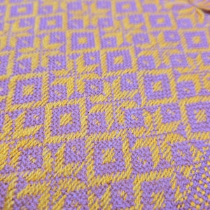 May include: A woven textile fabric featuring a repeating geometric pattern of interlocking squares and star-like shapes. The design is created with a vibrant combination of bright yellow and light purple threads, showing a dense, textured weave.