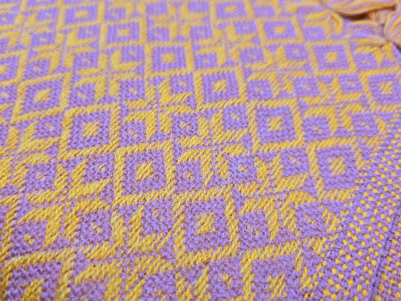 May include: A woven textile fabric featuring a repeating geometric pattern of interlocking squares and star-like shapes. The design is created with a vibrant combination of bright yellow and light purple threads, showing a dense, textured weave.