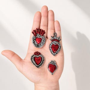 May include: Four heart-shaped charms with a metallic silver finish and a glossy red center. Each charm features intricate embossed detailing around the edges. Two of the charms include a decorative flame or arrow motif at the top, while two are plain.