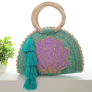Puede incluir: Bolso tejido turquesa y beige con diseño de concha. La concha es morada con detalles de cuentas doradas. El bolso tiene un asa redonda y borlas turquesas. Una pequeña planta en maceta está en el fondo.