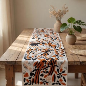 May include: A white fabric table runner featuring an intricate embroidered pattern of birds, leaves, and floral motifs in shades of burnt orange and black. The runner is laid flat across a wooden table, showcasing detailed stitching and organic shapes.