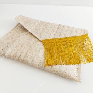 Op de afbeelding: Een rechthoekige clutch in envelopstijl gemaakt van geweven lichtbeige natuurlijke vezels. De flap is voorzien van een decoratieve rand van lange, levendige mosterdgele franjes die over de voorkant van de geweven tas hangen.