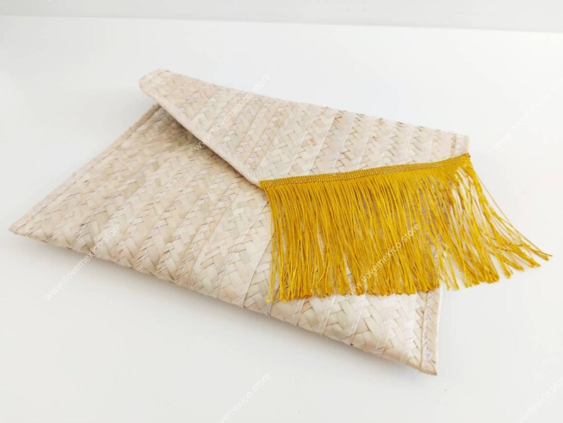 Op de afbeelding: Een rechthoekige clutch in envelopstijl gemaakt van geweven lichtbeige natuurlijke vezels. De flap is voorzien van een decoratieve rand van lange, levendige mosterdgele franjes die over de voorkant van de geweven tas hangen.