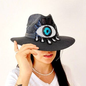 May include: A black fedora hat with a large blue eye sequin patch. The eye has a white iris and black eyelashes. The hat is being held by a person.