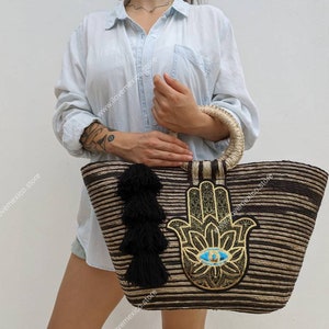 May include: A large, woven tote bag with a brown and beige striped pattern. The bag features a large, embroidered hamsa hand with a blue eye in the centre. The bag has a black tassel hanging from the side.