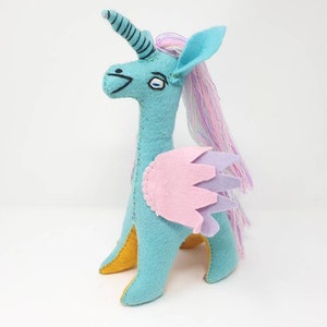 Puede incluir: Un peluche de unicornio de fieltro turquesa con un cuerno en espiral, una larga melena de hilo rosa y morado, y alas superpuestas en rosa claro y lavanda. La criatura tiene detalles bordados en ojos y boca, con tela amarilla visible en las patas.