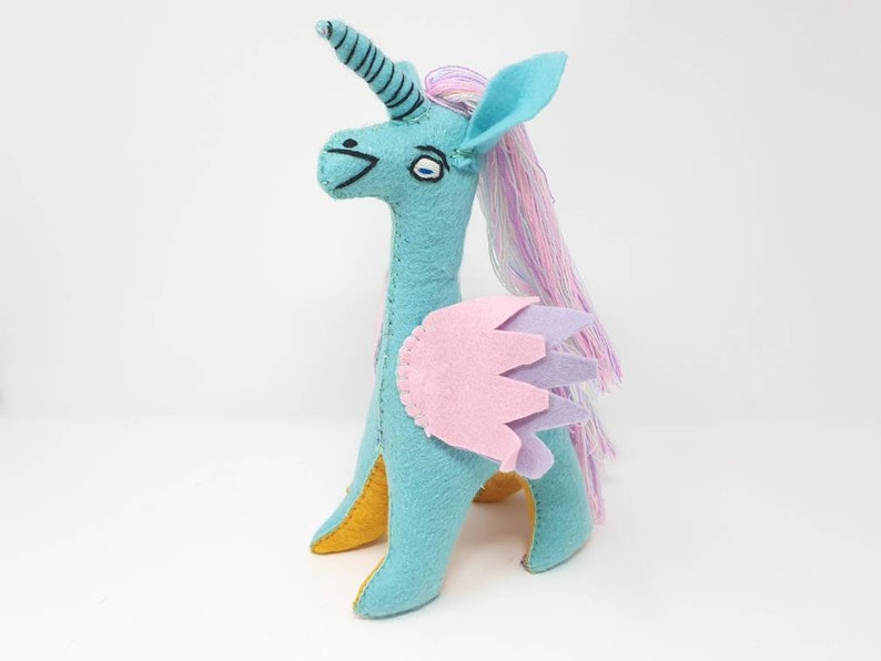 Puede incluir: Un peluche de unicornio de fieltro turquesa con un cuerno en espiral, una larga melena de hilo rosa y morado, y alas superpuestas en rosa claro y lavanda. La criatura tiene detalles bordados en ojos y boca, con tela amarilla visible en las patas.