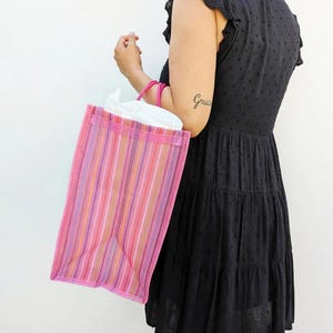 May include: A rectangular tote bag featuring vertical stripes in shades of pink, purple, orange, and white. The bag has two pink circular handles and is constructed from a semi-transparent material, revealing a white interior lining inside.